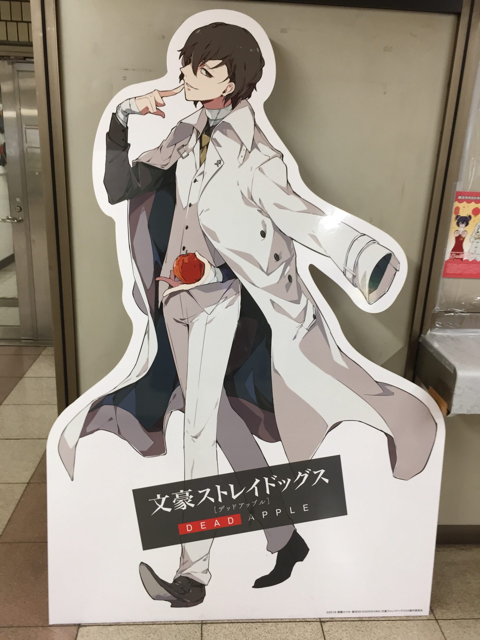 横浜桜木町駅に設置された太宰治の等身大キャラクターパネル。太宰はDEAD APPLEの描き下ろしデザインの姿で、白いツリーピーススーツを纏っている。左手が赤いリンゴを持っている。右手の人差し指が唇にあたっている。