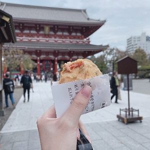 背景に浅草寺があり、屋台で買ったスナックとそれを持つ手が手前に写っている