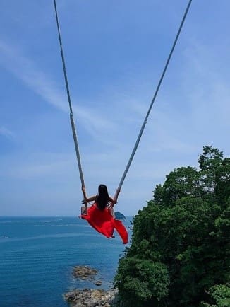 海と空に向かって赤いドレスを着た女の人がブランコを漕いでいる