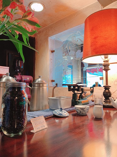 カフェのテーブル。赤いランプシェードのランプや、銀製のポット、コーヒー豆などが置かれている。
