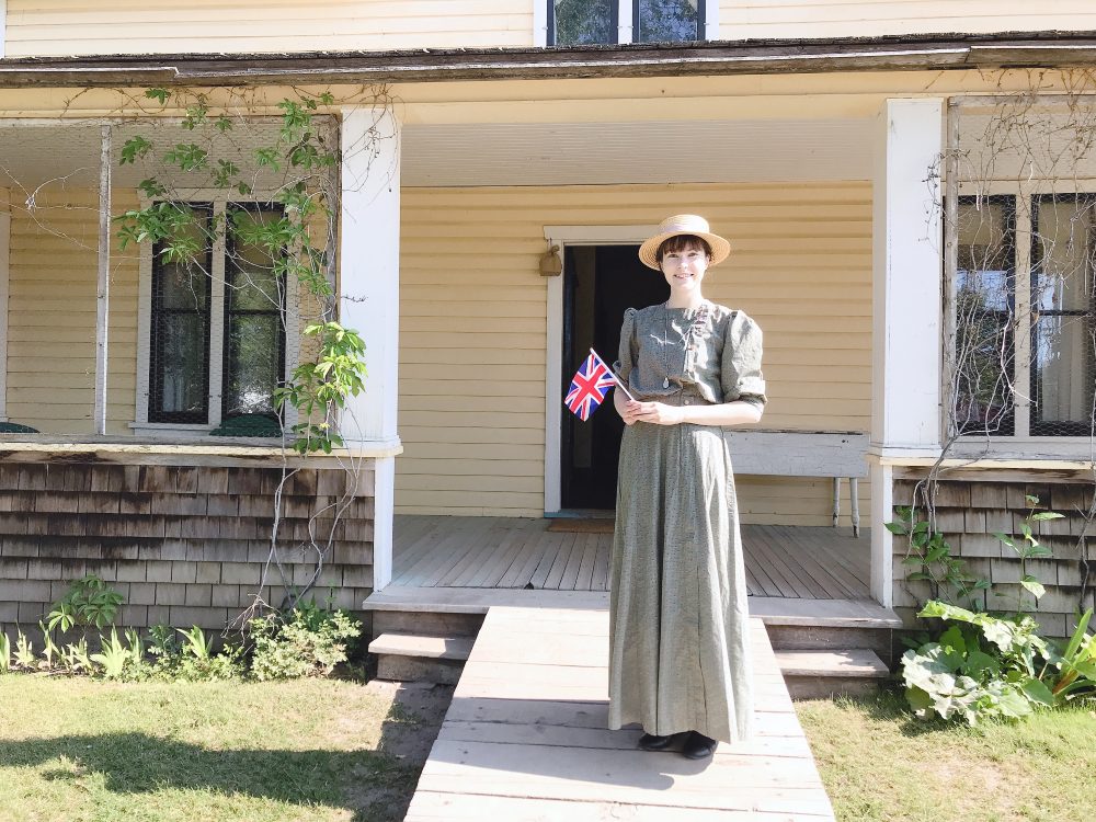 1910年代の服装(長いパフスリーブのある緑色ワンピースとかんかん帽)をしているラターナスはイギリスの国旗を手に持ち、黄色い牧場の家の前に立っています。