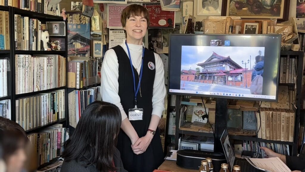 元気に挨拶をする弊社スタッフのジェイリーン。斜陽館の生中継が後ろのモニターに写っている。