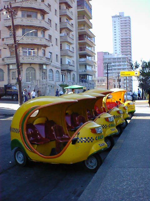 ココナッツのような形をした黄色い三輪車バイクタクシーが五台道の脇に駐車している。背景にはコロニアル様式の高層アパートが見える。