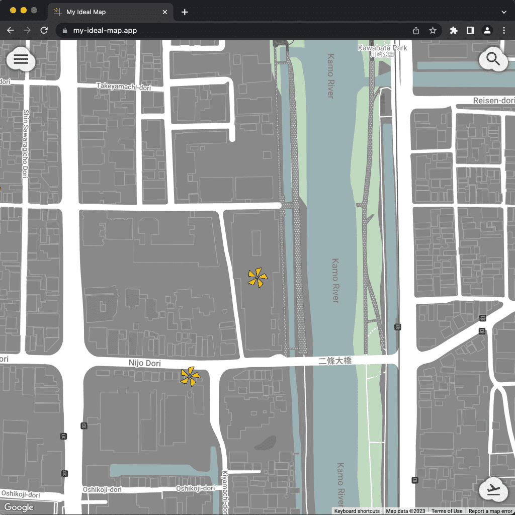 道路は白、建物が建つ場所は灰色で示されている地図。中央から右にずれた位置に、Kamo River と表示されている暗めの水色のエリアが上から下へ貫いている。中央とその左下あたりに、山吹色の5枚の花びらからなる花のようなアイコンが表示されている。雲のような形をした白いボタンが三つ。一つは左上の角にあり、横三本線のメニューアイコンが、もう一つは右上の角で虫眼鏡アイコン、最後の一つは右下の角で離陸する飛行機のアイコン、が描かれている。最上部にはブラウザーのアドレスバーがあり、そこには、"my-ideal-map.app" と表示されている。