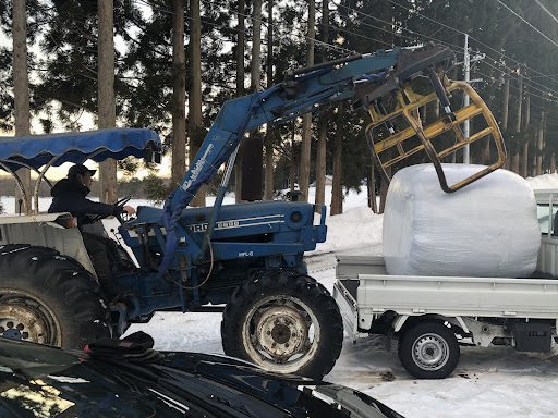 軽トラから餌のロールを下すトラクターのような機械。アームの先端でロールをつまんで持ち上げる。