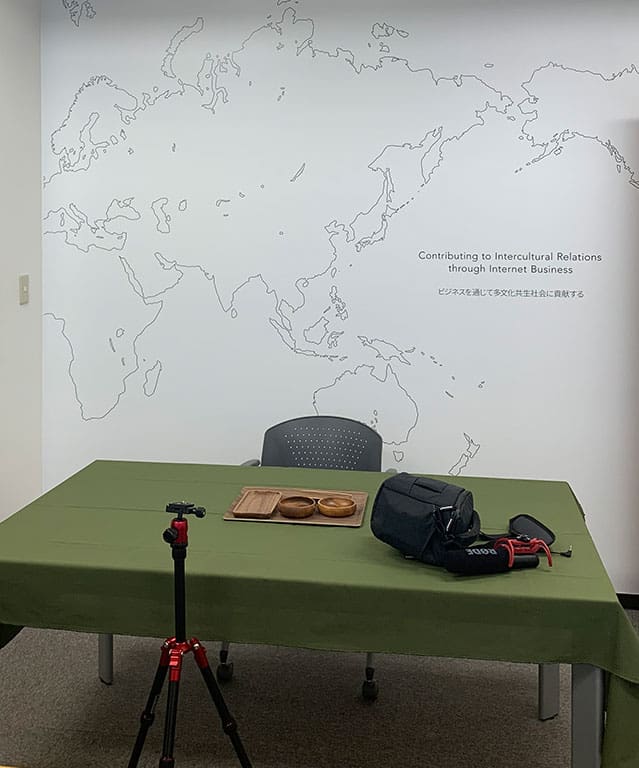 撮影場所は東京オフィスで白い壁の前に緑のテーブルクロスをひいたテーブルがあり、撮影機材がおかれています。